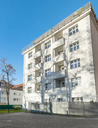 Mehrstöckiges Wohnhaus mit Balkonen und mehreren Fenstern, umgeben von Bäumen und Gehweg.