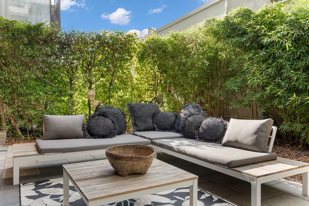 Moderne Terrasse mit Sofaecke, grauen Kissen und Holz-Couchtisch, umgeben von grünen Pflanzen.