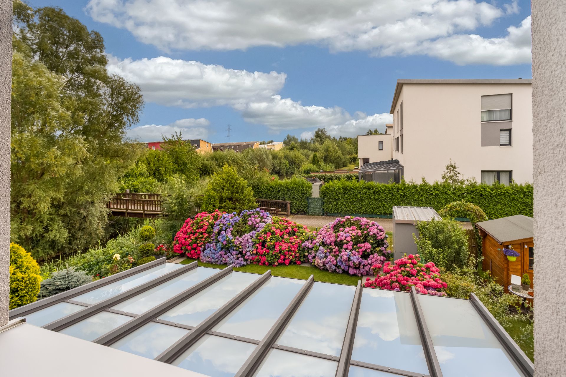 Blick auf Garten mit bunten Blumen, Glasdach, Bäumen und Wohngebäuden im Hintergrund.