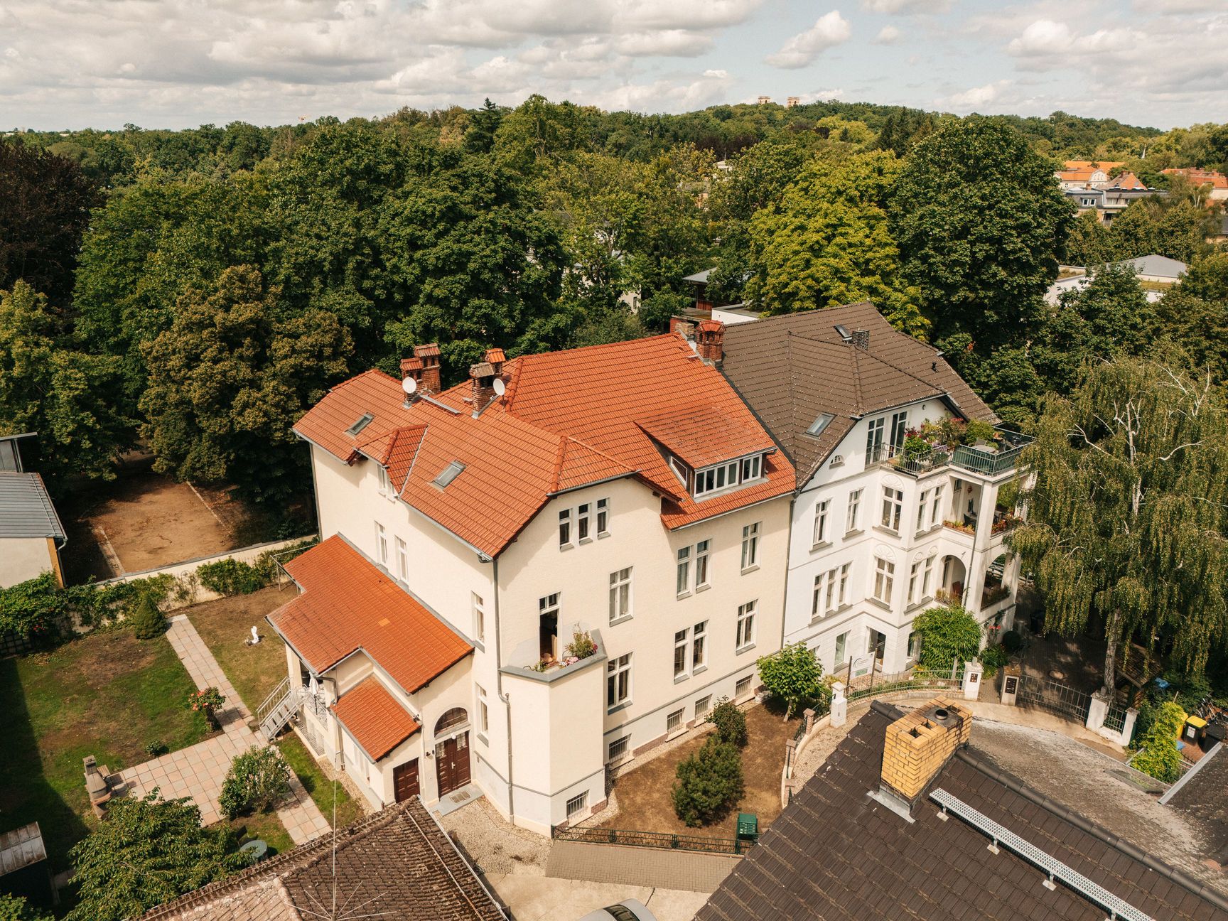 Großes Wohnhaus mit rotem Dach, umgeben von Bäumen und Gartenanlage.