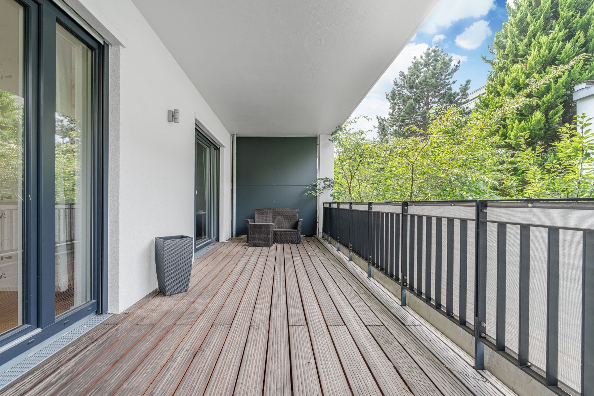 Großer Balkon mit Holzdielen, dunklem Geländer, Sitzmöbel und Blick auf grüne Bäume.