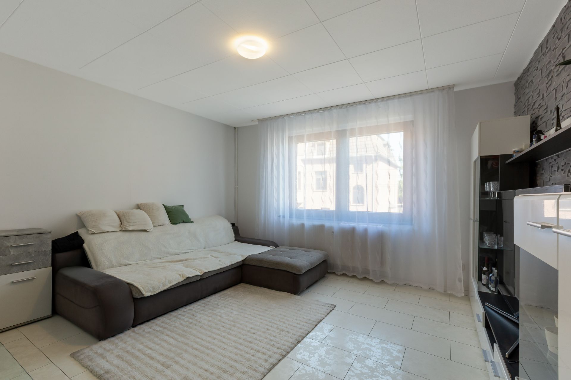 Wohnzimmer mit Sofa, Teppich, großem Fenster mit Vorhängen und Wandregal.