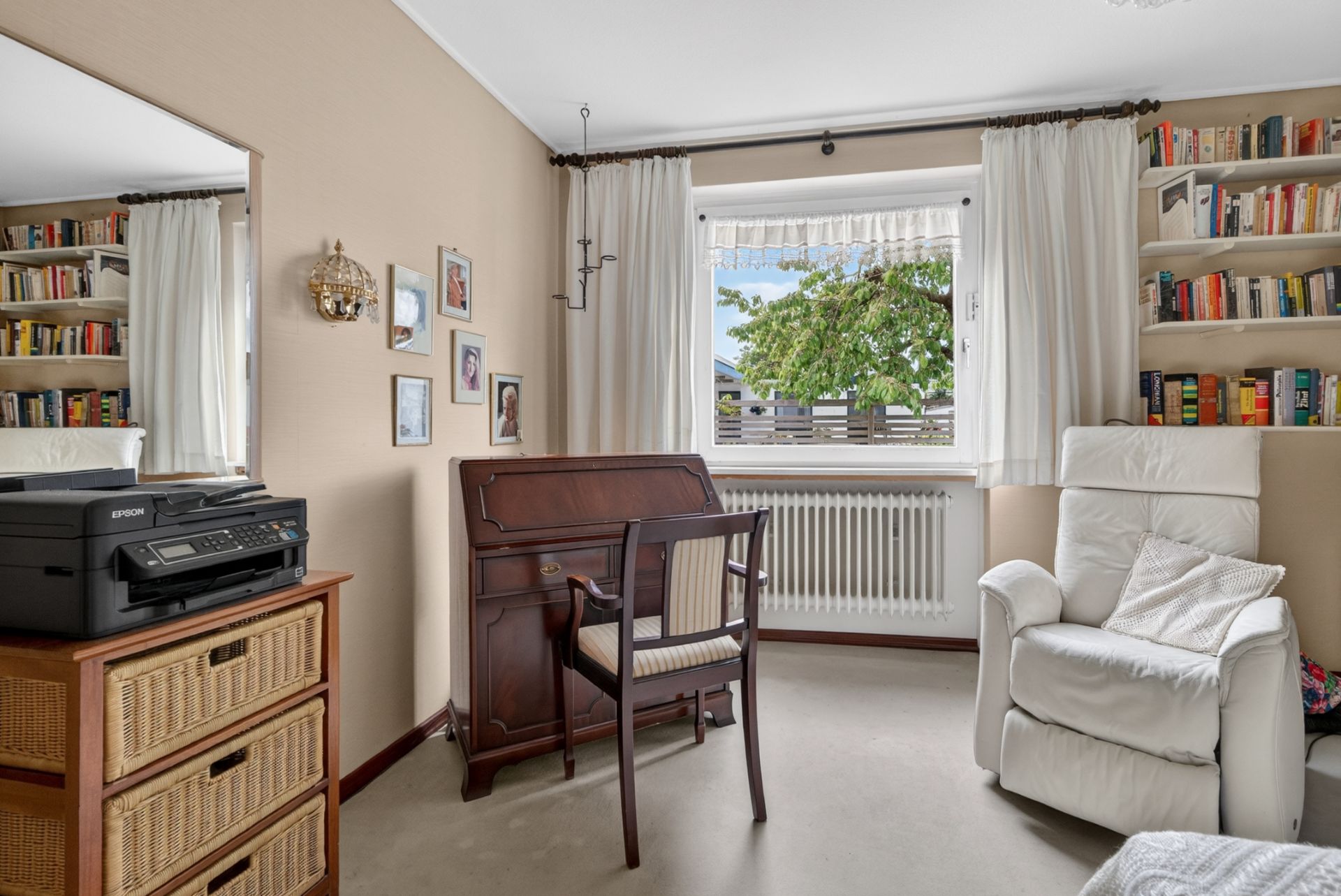 Helles Arbeitszimmer mit Sessel, Schreibtisch, Bücherregalen, Drucker und Fenster mit Blick ins Grüne.