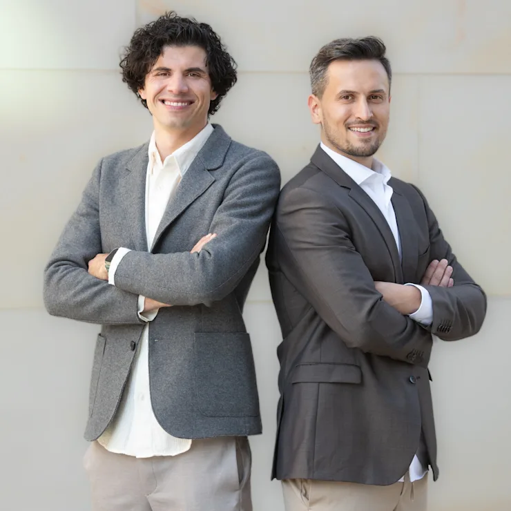 Zwei Männer stehen nebeneinander vor einer hellen Wand und lächeln in die Kamera.