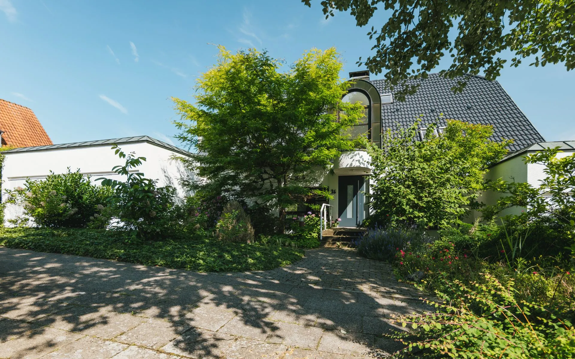 Modernes Wohnhaus mit dunklem Dach, von Bäumen und Sträuchern umgeben, vor einem gepflasterten Weg.