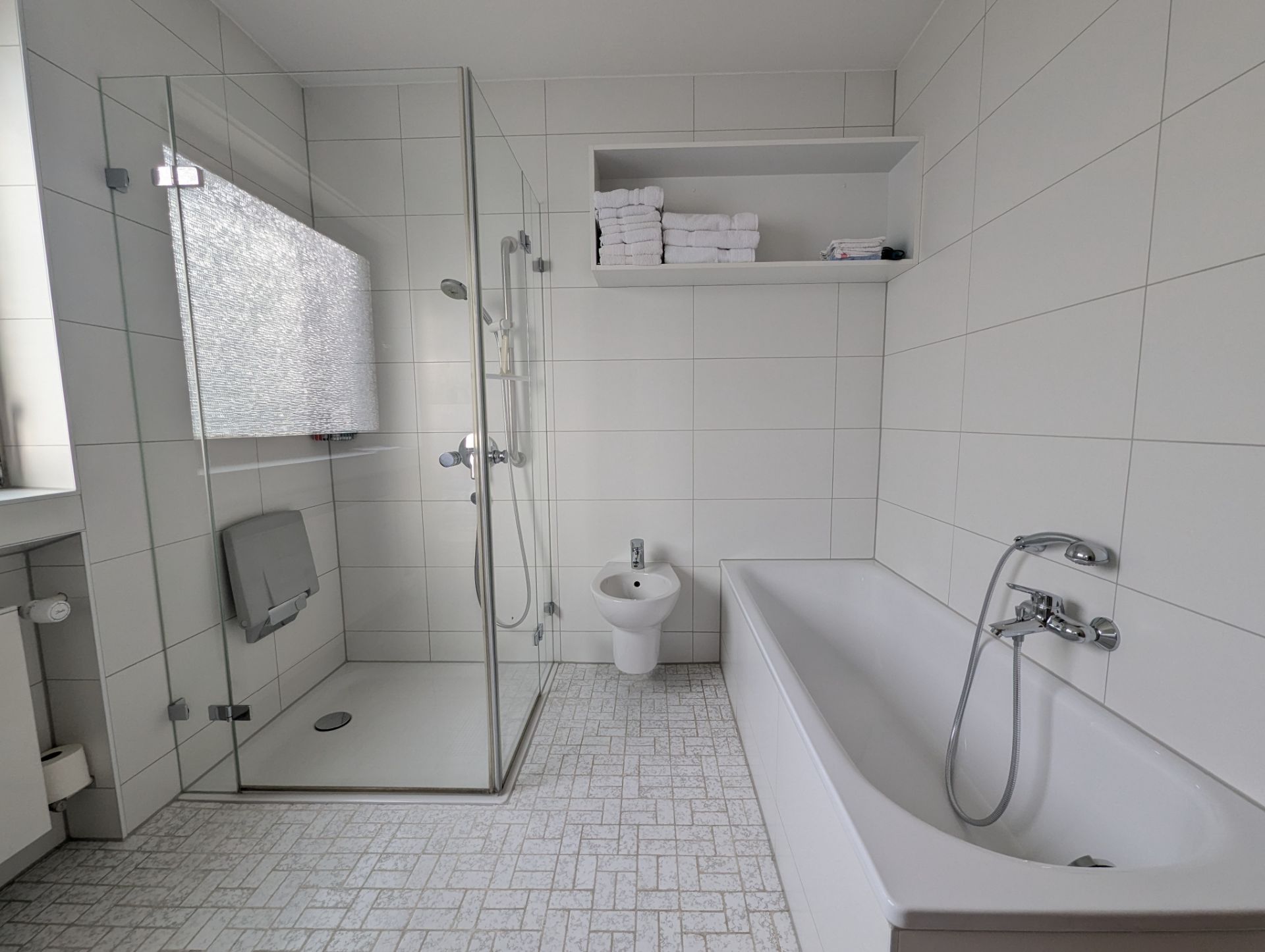 Modernes Bad mit Dusche, Badewanne, Bidet und weißen Fliesen. Regale mit Handtüchern an der Wand.