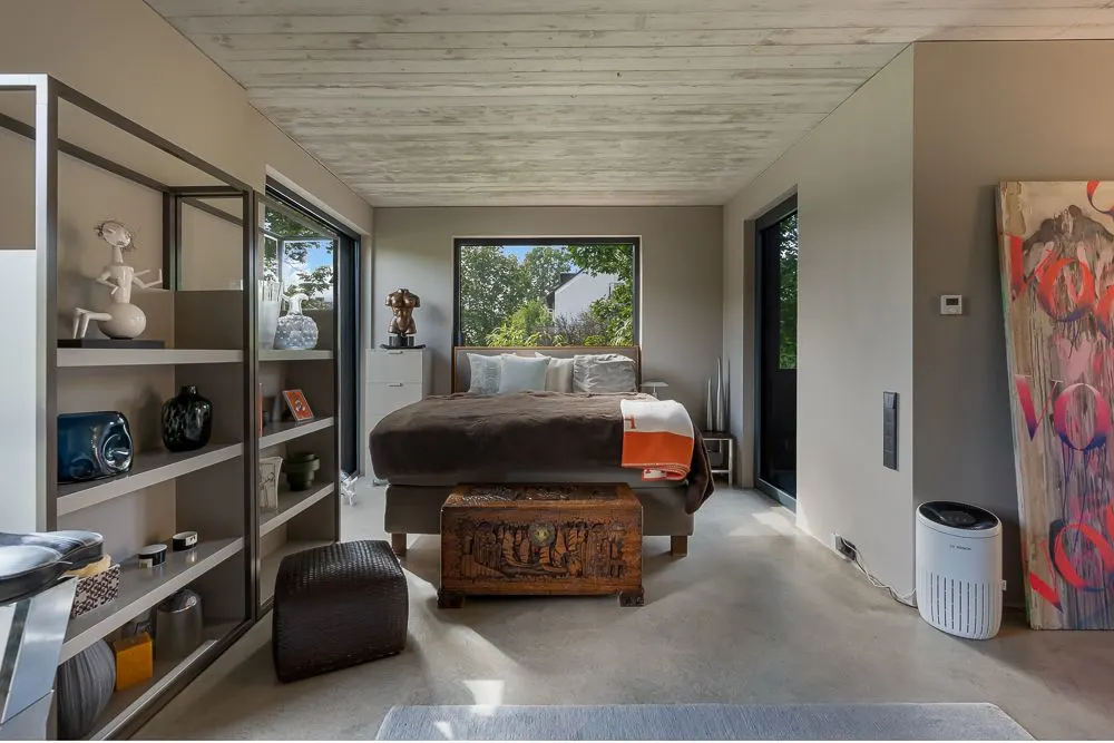 Modernes Schlafzimmer mit großem Fenster, Doppelbett, Regal, Truhe und Kunstwerk an der Wand.