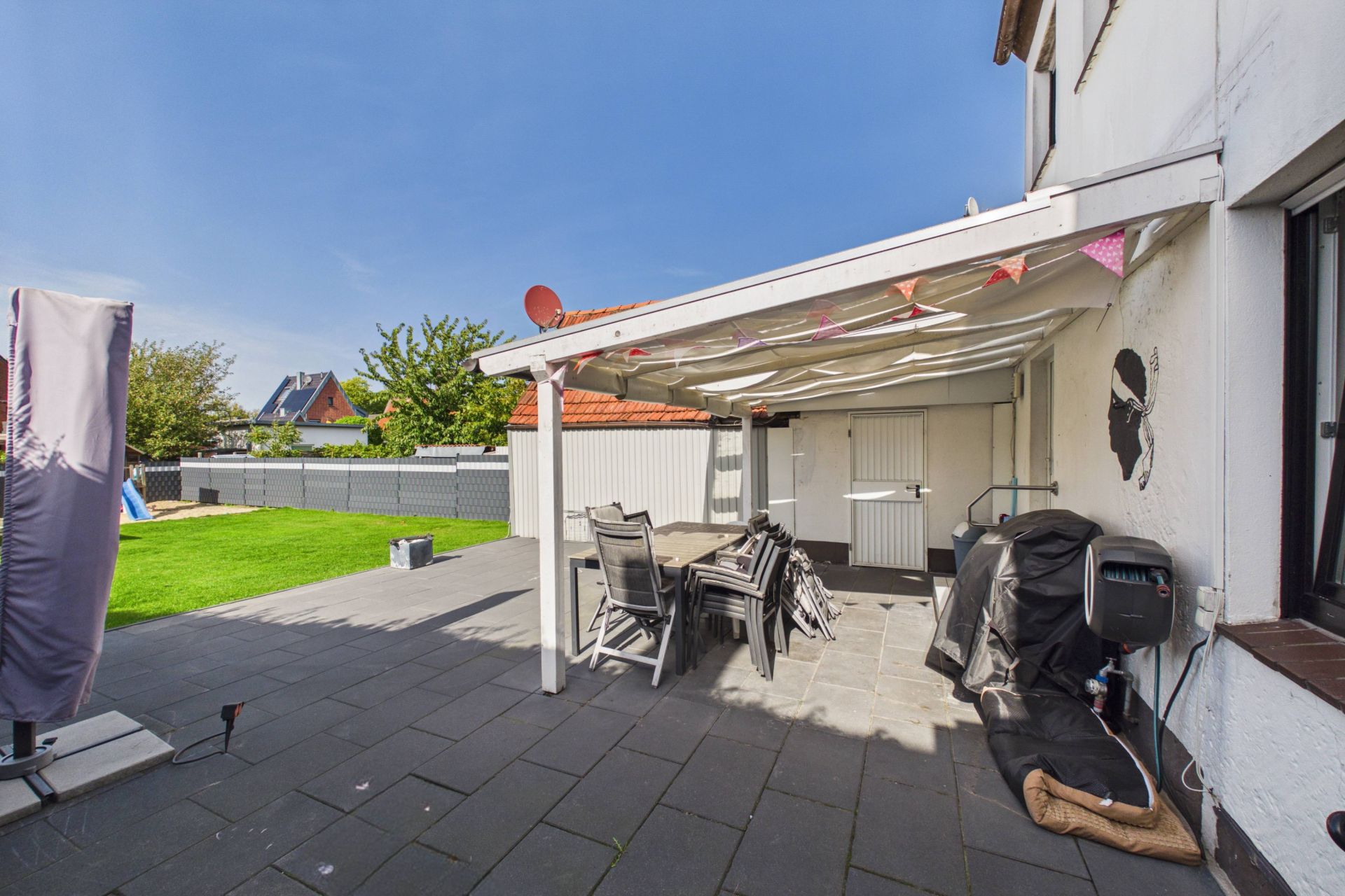 Überdachte Terrasse mit Gartentisch, Stühlen und Blick auf Garten mit Rasen und Zaun.