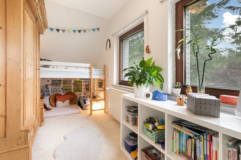 Helles Kinderzimmer mit Hochbett, Regalen, Büchern, Pflanzen und großen Fenstern.