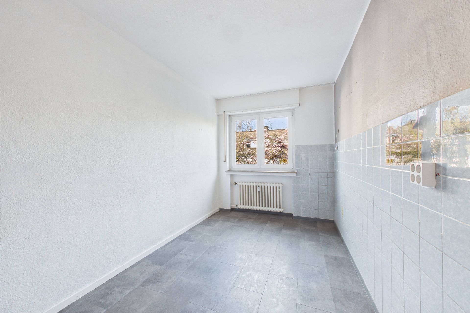 Leerer, schmaler Raum mit Fenster, Heizkörper und teils gefliester Wand.