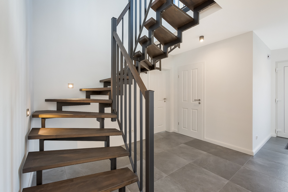 Moderne Holztreppe in einem hellen Flur mit grauen Bodenfliesen und weißen Türen.