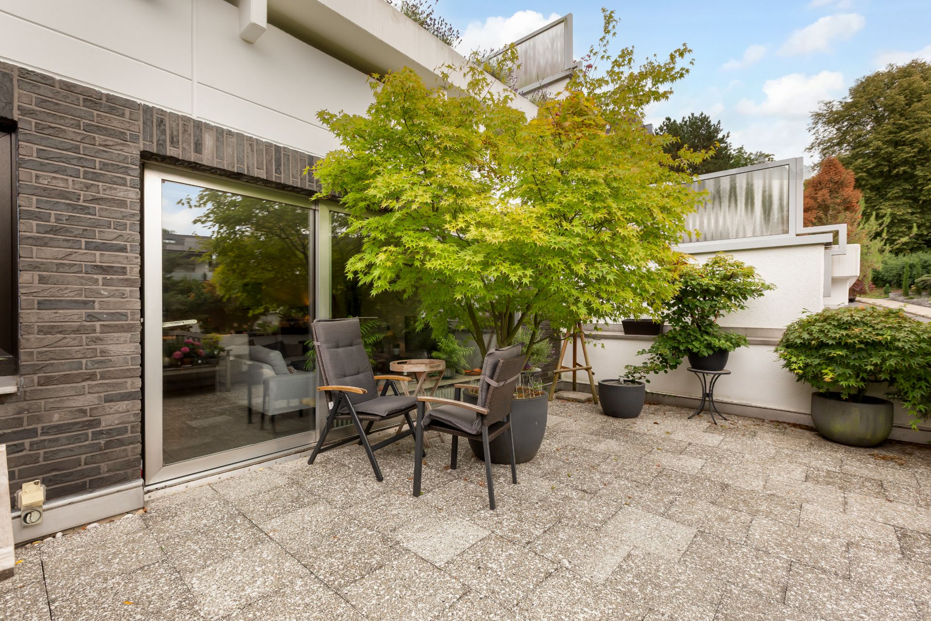 Große Terrasse mit Gartenmöbeln, mehreren Pflanzenkübeln und einem Baum neben dem Haus.