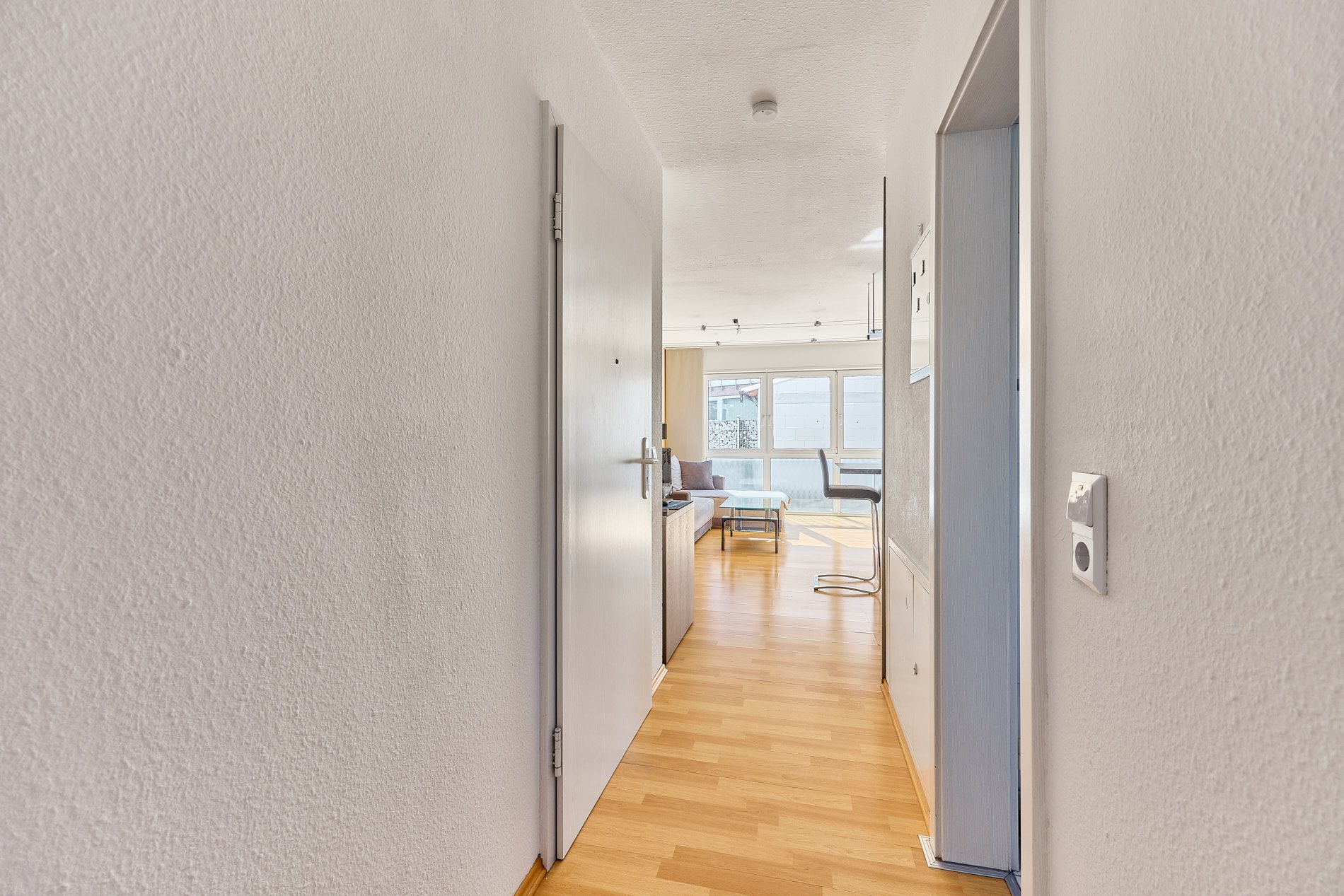 Heller Flur mit Holzfußboden, Blick ins Wohnzimmer mit Sofa und großen Fenstern.