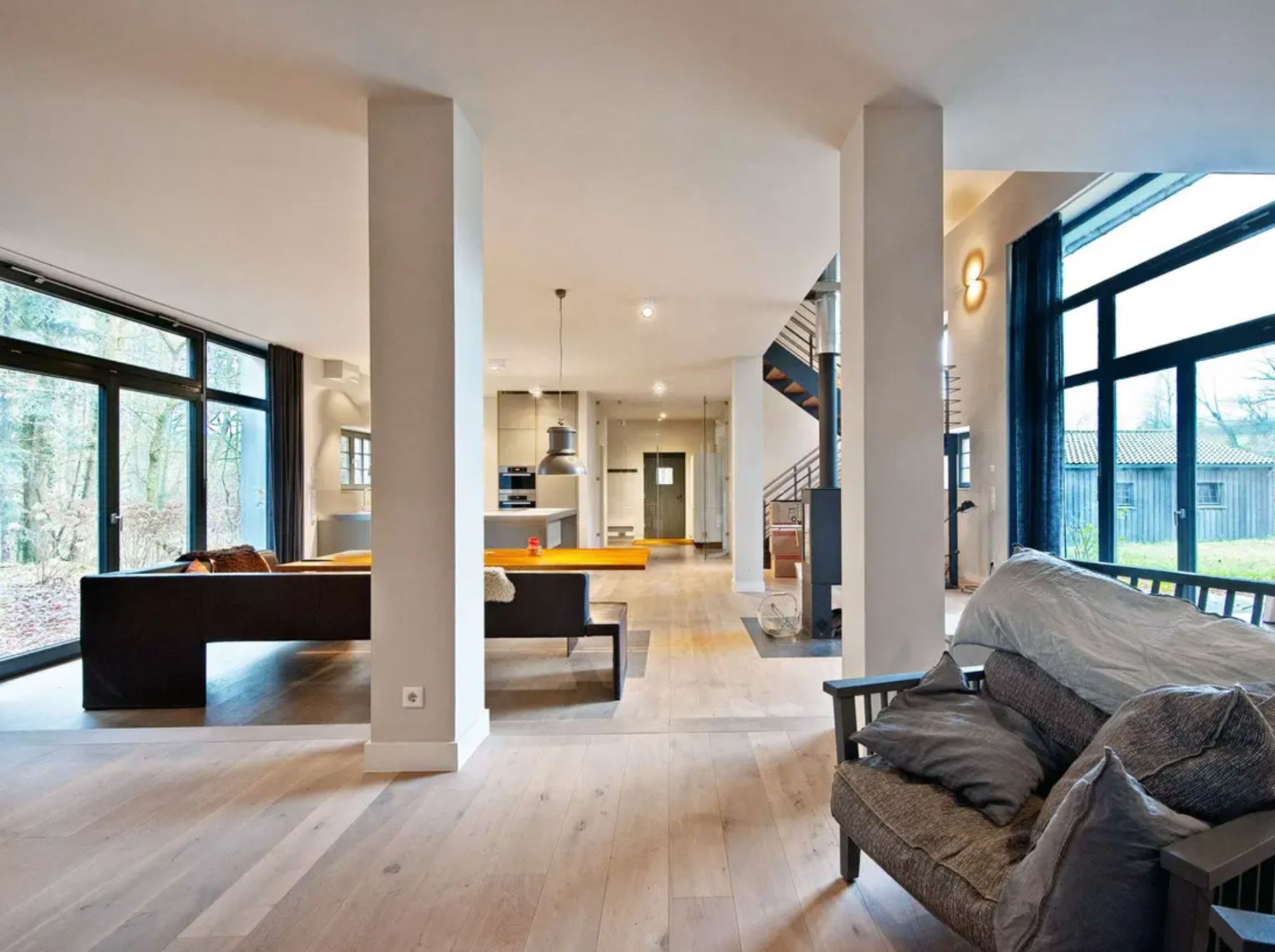 Offenes Wohnzimmer mit großen Fenstern, Sofas, Holzboden und Blick auf eine Treppe.