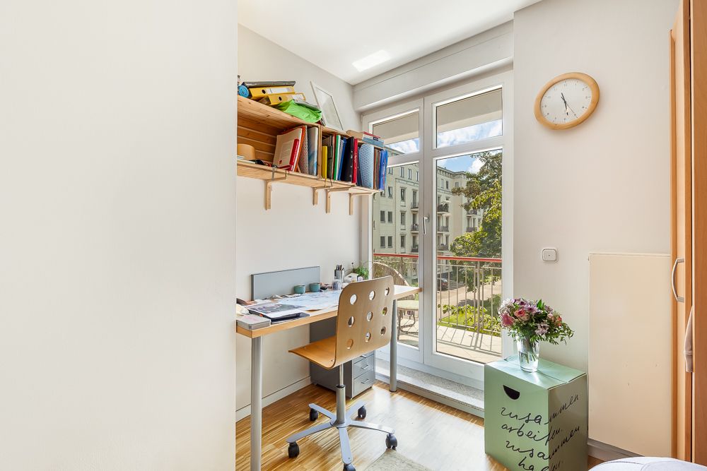Helles Arbeitszimmer mit Schreibtisch, Regal, Stuhl und Blumen, großer Fensterblick auf Wohnhäuser.