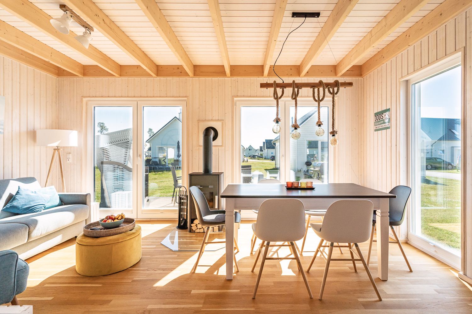 Helles Wohnzimmer mit Esstisch, vier Stühlen, Sofa und Holzofen, große Fenster mit Blick nach draußen.