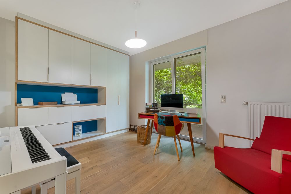 Heller Arbeitsraum mit Schreibtisch, Regalwand, rotem Sessel und E-Piano am Fenster.