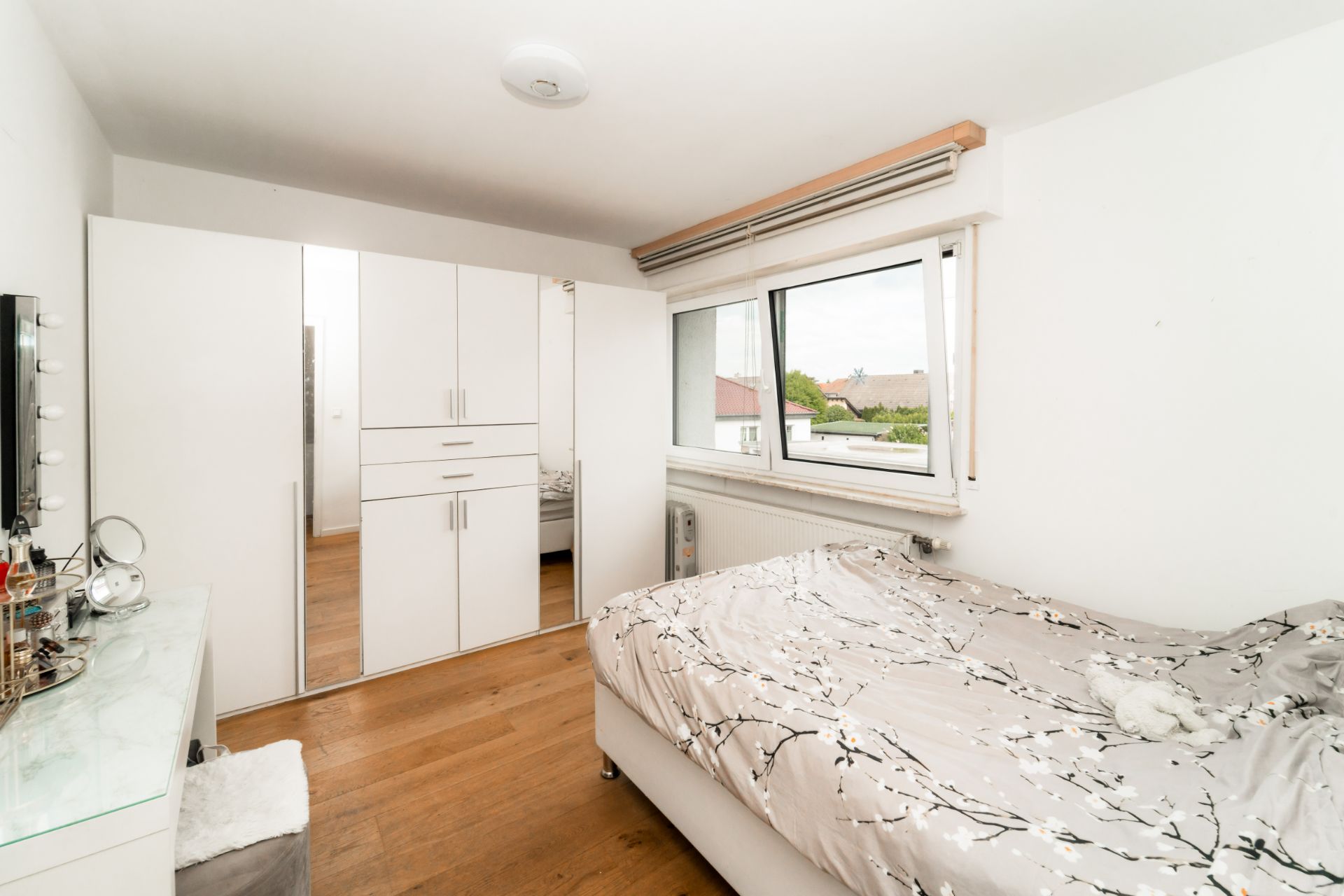 Helles Schlafzimmer mit Doppelbett, Kleiderschrank und großem Fenster auf Holzfußboden.