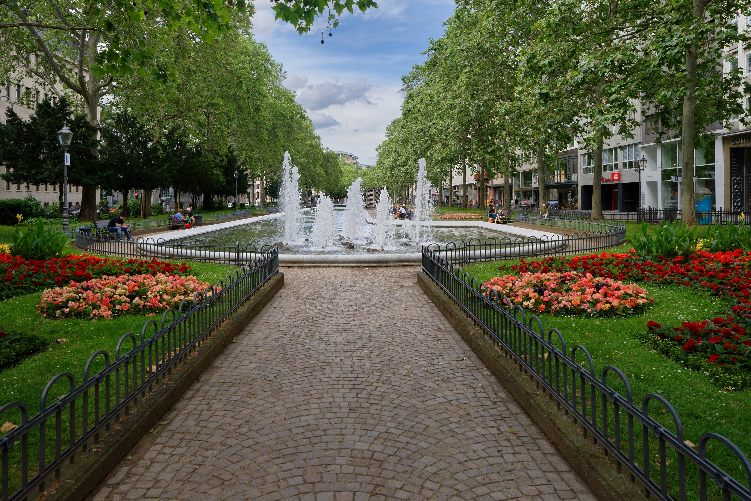 Gepflasterter Weg mit Blumenbeeten und Springbrunnen in einer grünen Parkanlage.