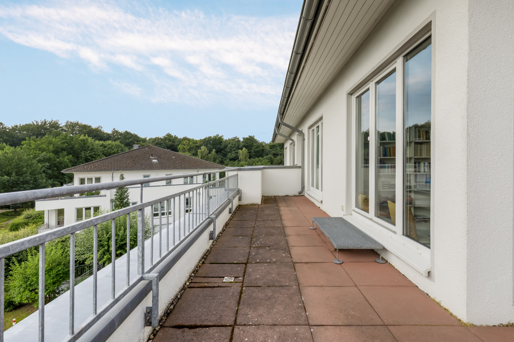 Großer Balkon mit Fliesenboden, Geländer und Blick auf Bäume und ein Nachbargebäude.