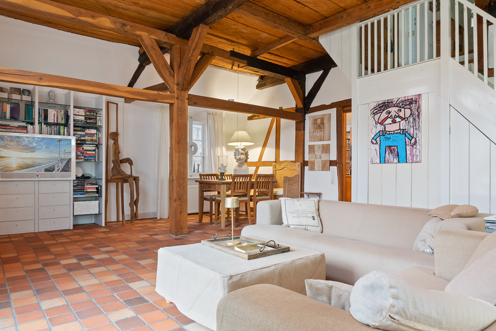 Helles Wohnzimmer mit Holzbalken, Sofa, Bücherregal und Esstisch im Hintergrund.