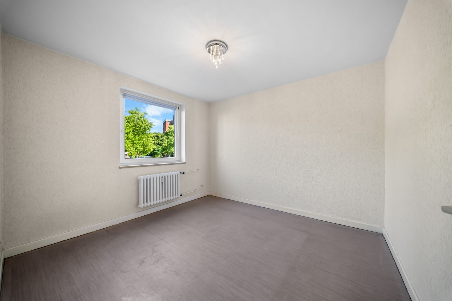 Leerer Raum mit Fenster, Heizkörper und Deckenleuchte, helle Wände, grauer Boden.