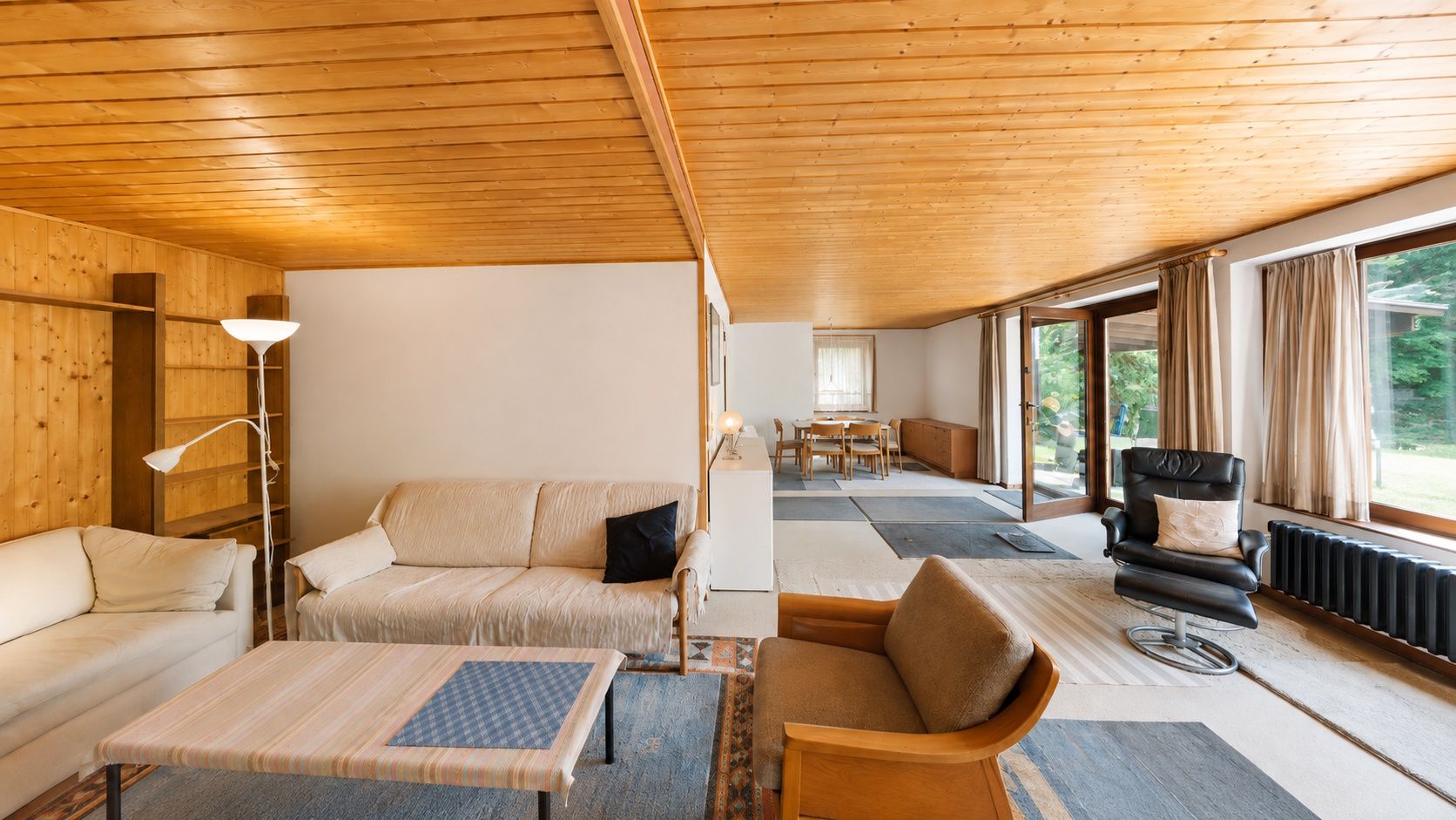 Wohnzimmer mit Holzboden und -decke, Sofas, Sessel, Couchtisch und großen Fenstern zum Garten.