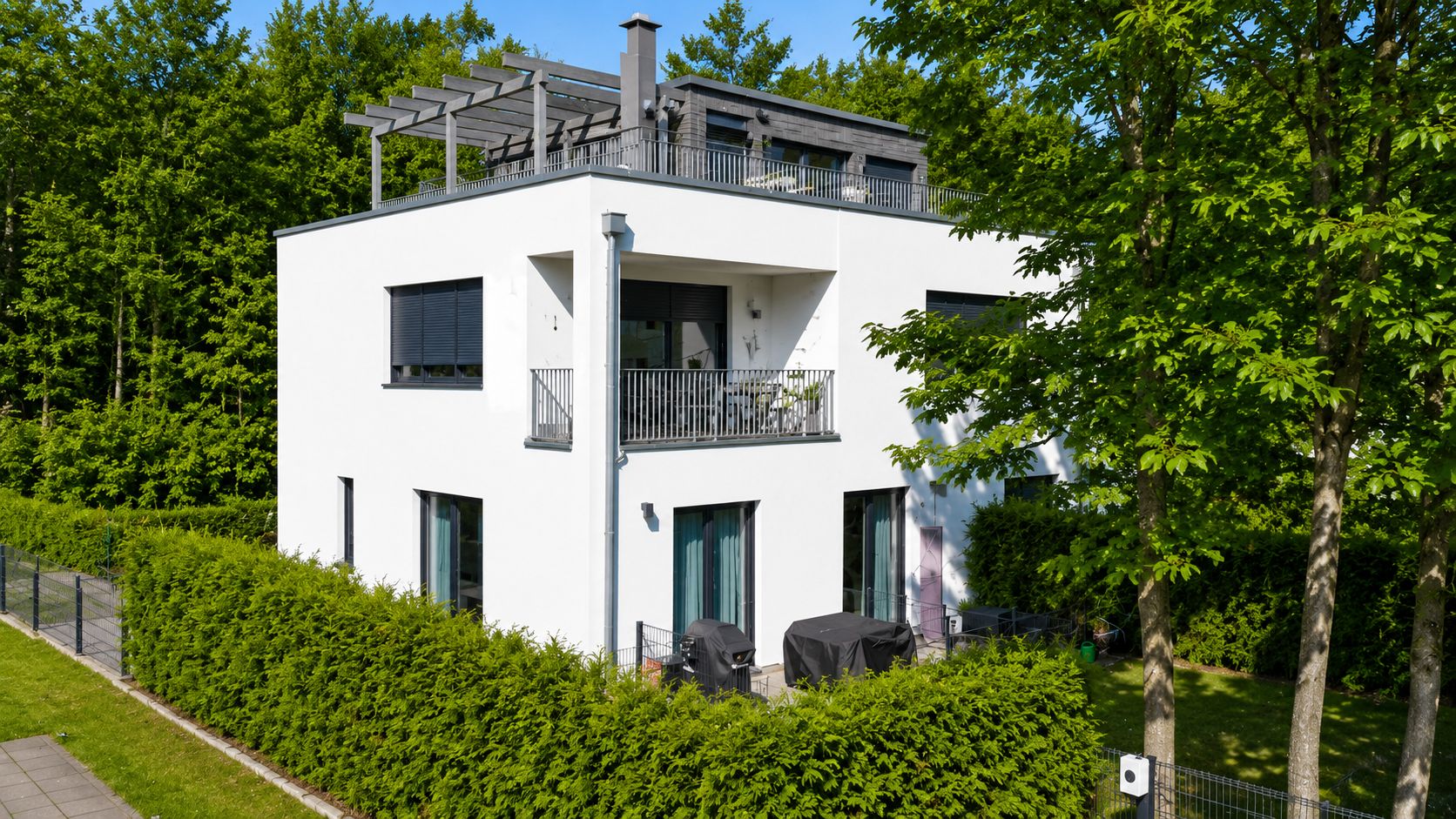 Modernes, weißes Wohnhaus mit Balkon, Terrasse auf dem Dach und umgeben von Bäumen und Hecken.