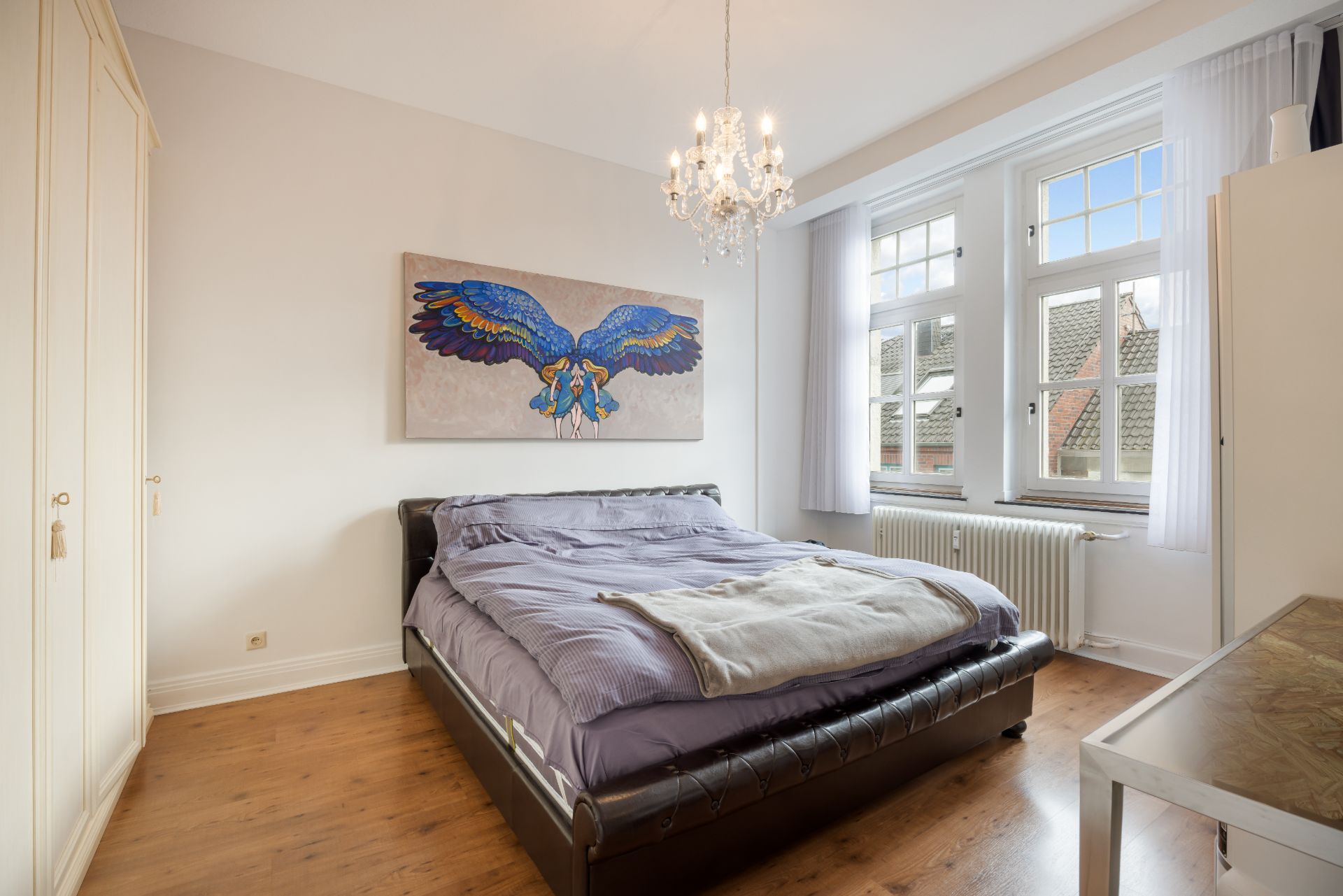 Helles Schlafzimmer mit Doppelbett, Kleiderschrank, Kronleuchter und großem Fenster.