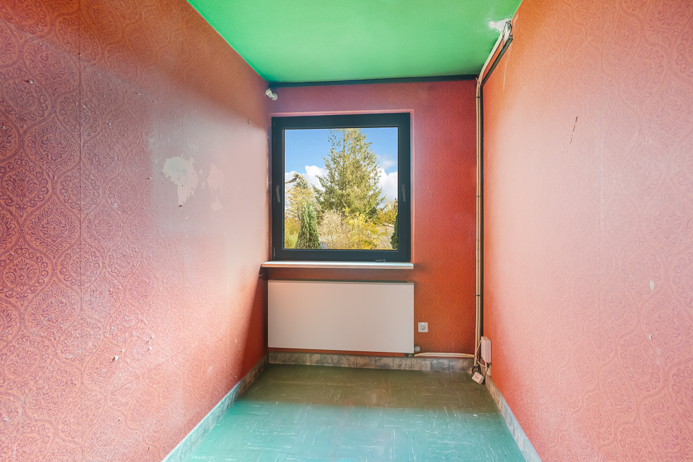 Kleiner leerer Raum mit roter Tapete, grünem Boden und Fenster mit Blick ins Grüne.