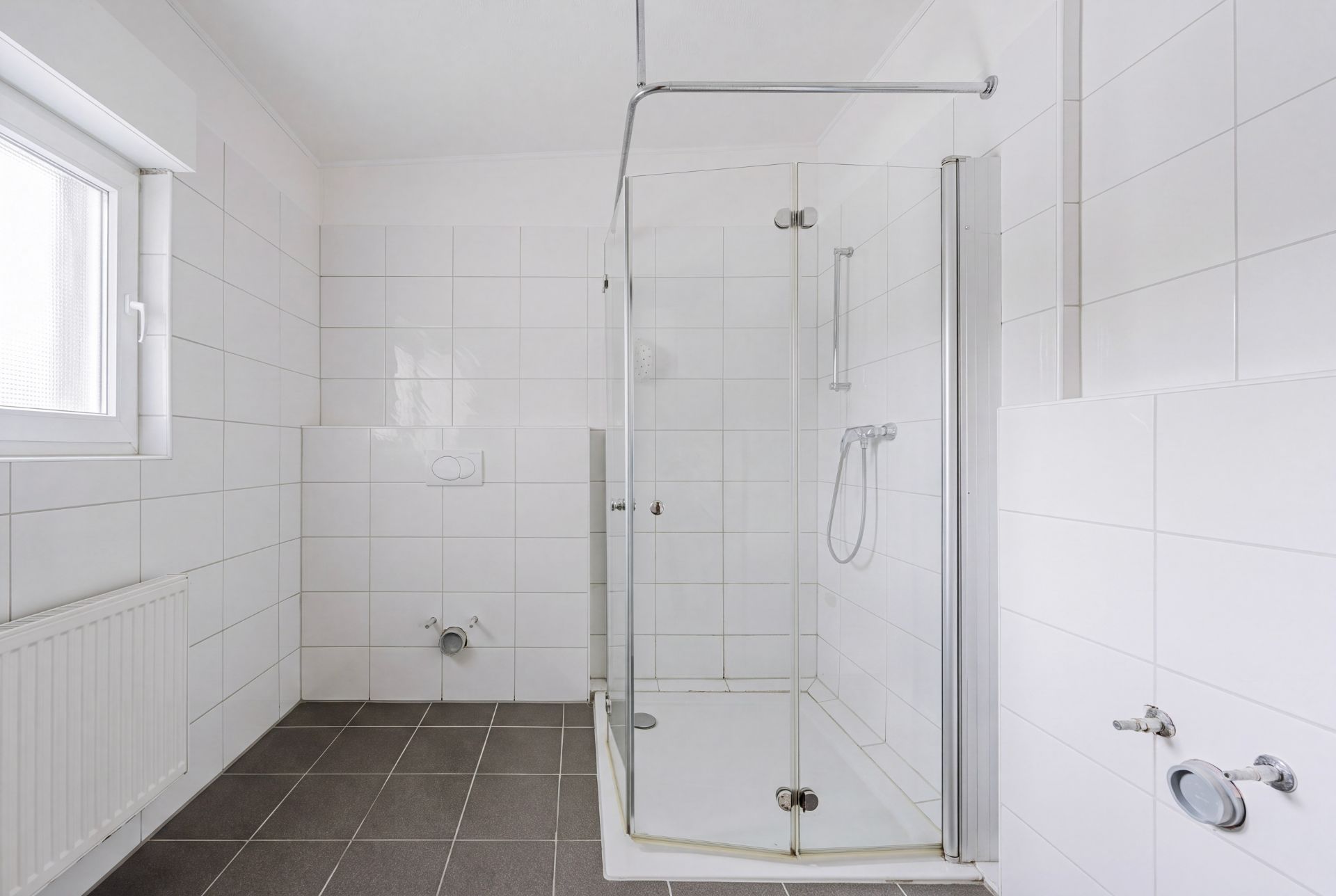 Modernes, weiß gefliestes Bad mit Dusche, Fenster und leerem Anschluss für Toilette.