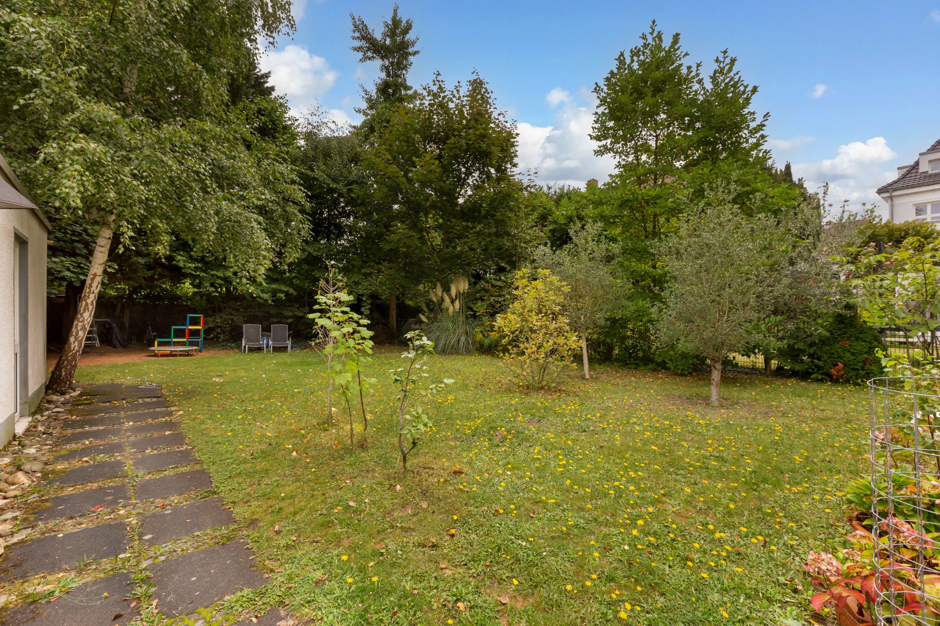 Großer Garten mit Rasen, Bäumen, Gartenweg und zwei Gartenstühlen im Hintergrund.