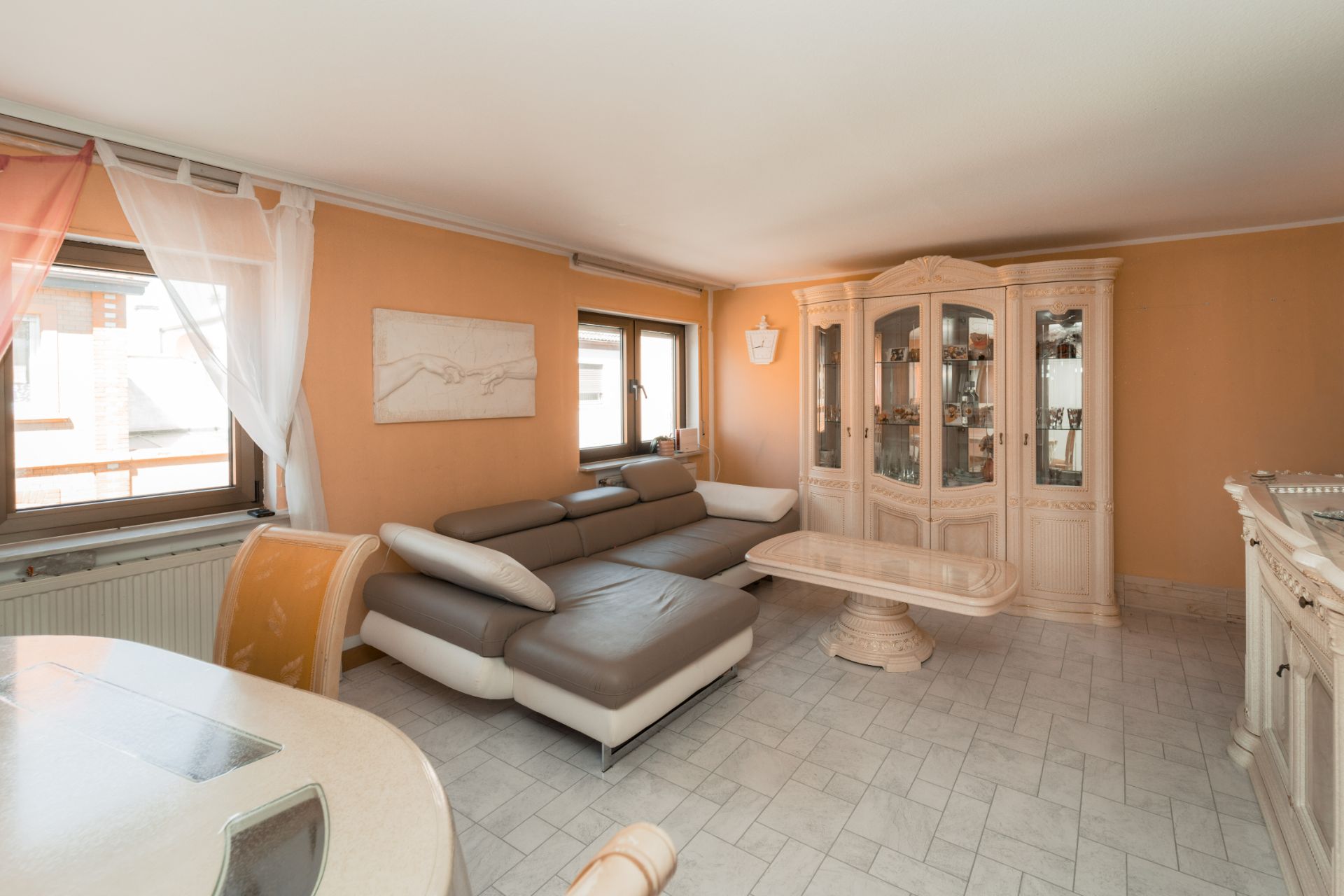 Wohnzimmer mit großer Eckcouch, Glastisch und hellem Vitrinenschrank vor orangefarbener Wand.