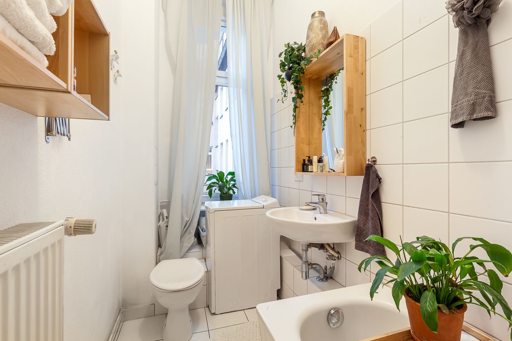 Helles Badezimmer mit Waschbecken, Toilette, Waschmaschine, Spiegel, Pflanzen und weiß gefliesten Wänden.
