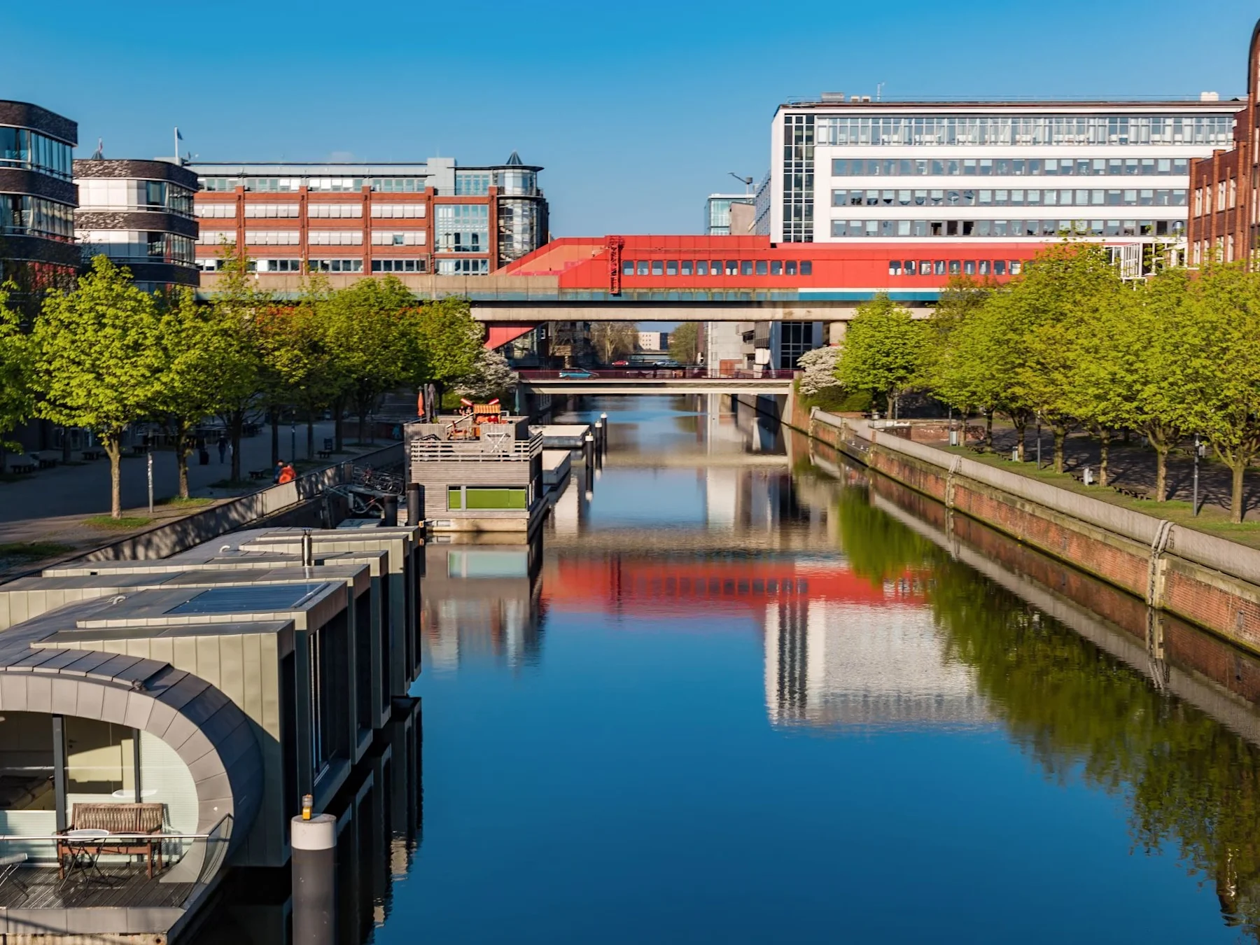 Moderne Hausboote am ruhigen Kanal, flankiert von Bäumen und Bürogebäuden bei klarem Himmel.