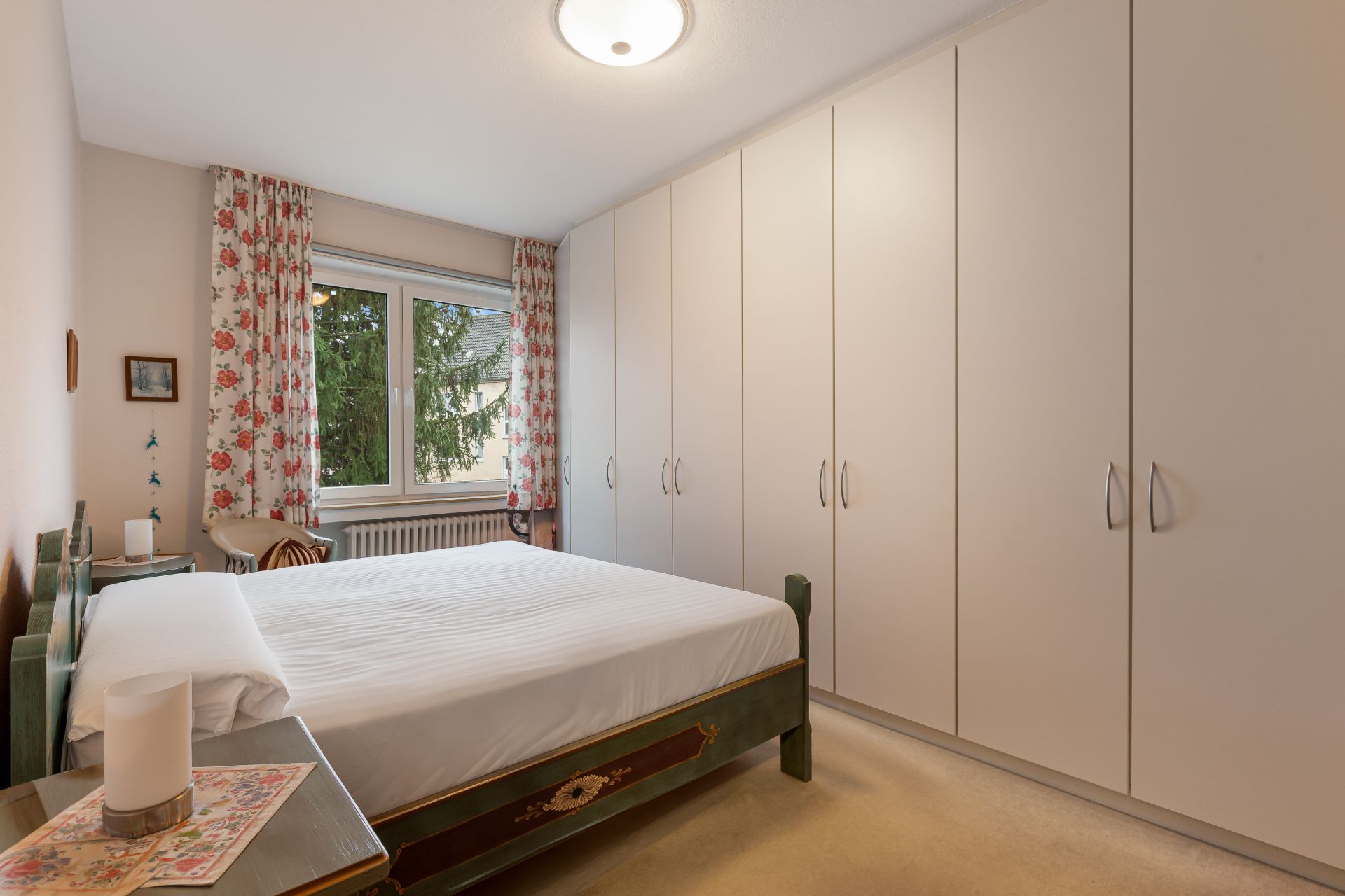 Schlafzimmer mit großem weißen Kleiderschrank, Doppelbett und Fenster mit Blumenvorhängen.