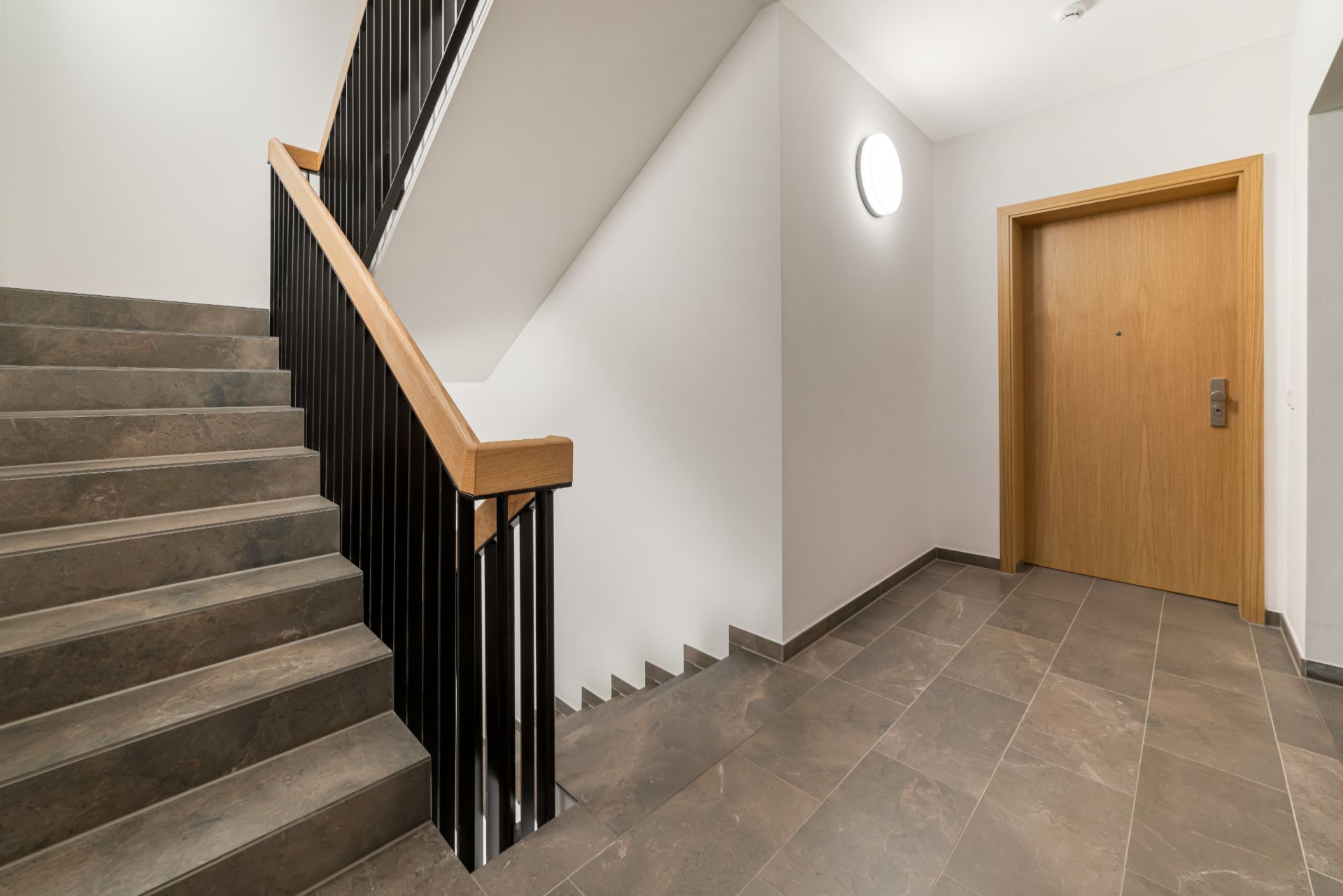 Treppenhaus mit braunen Fliesen, Geländer und einer Holztür auf der rechten Seite.