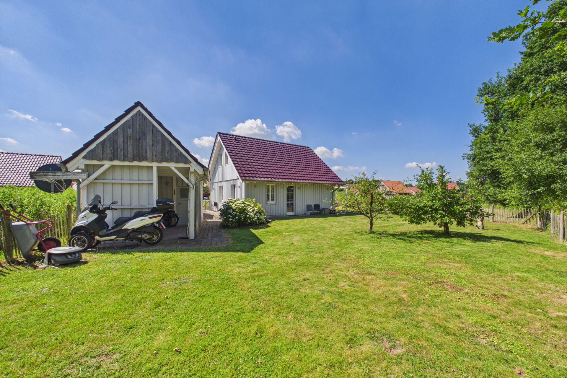 Haus mit rotem Dach und Nebengebäude auf großer, grüner Wiese, Motorroller unter Carport.