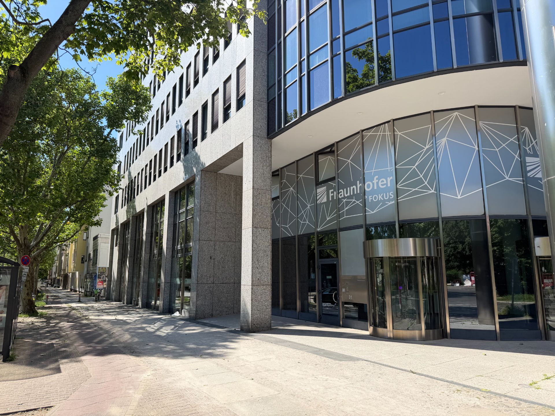 Moderne Glasfassade eines Bürogebäudes mit großem Eingangsbereich und Bäumen am Gehweg.