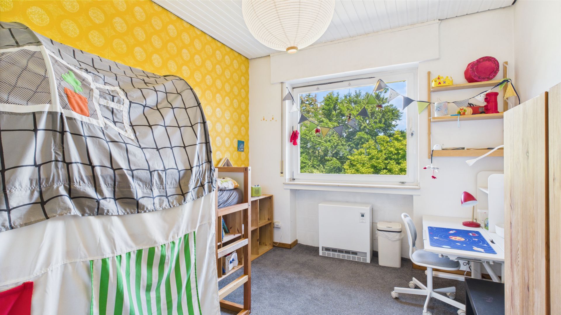Helles Kinderzimmer mit Spielbett, Schreibtisch und großem Fenster mit Blick ins Grüne.