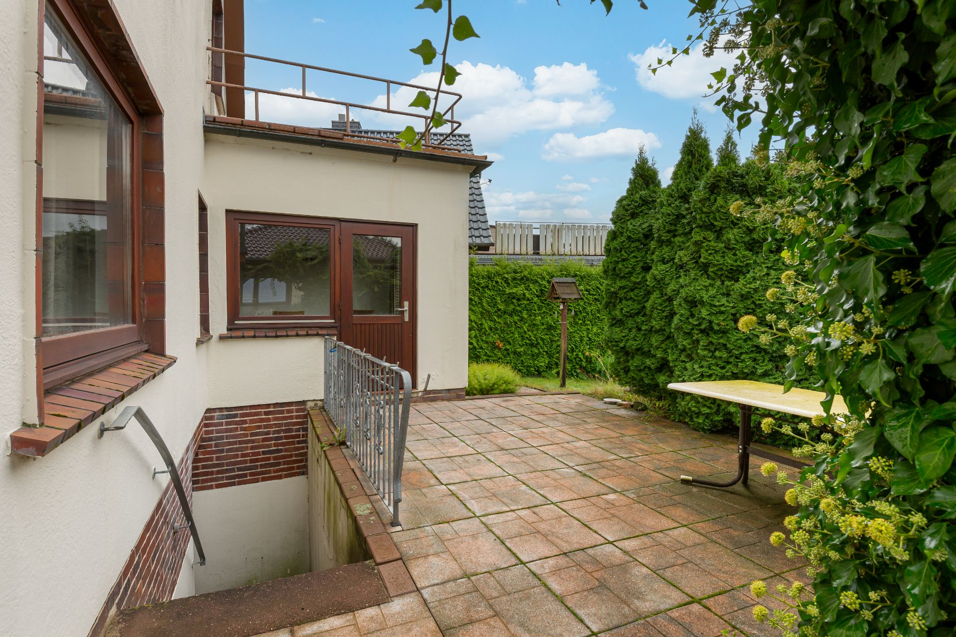 Gepflasterte Terrasse mit Tisch, umgeben von grüner Hecke und Zugang zu einem Kellerabgang.