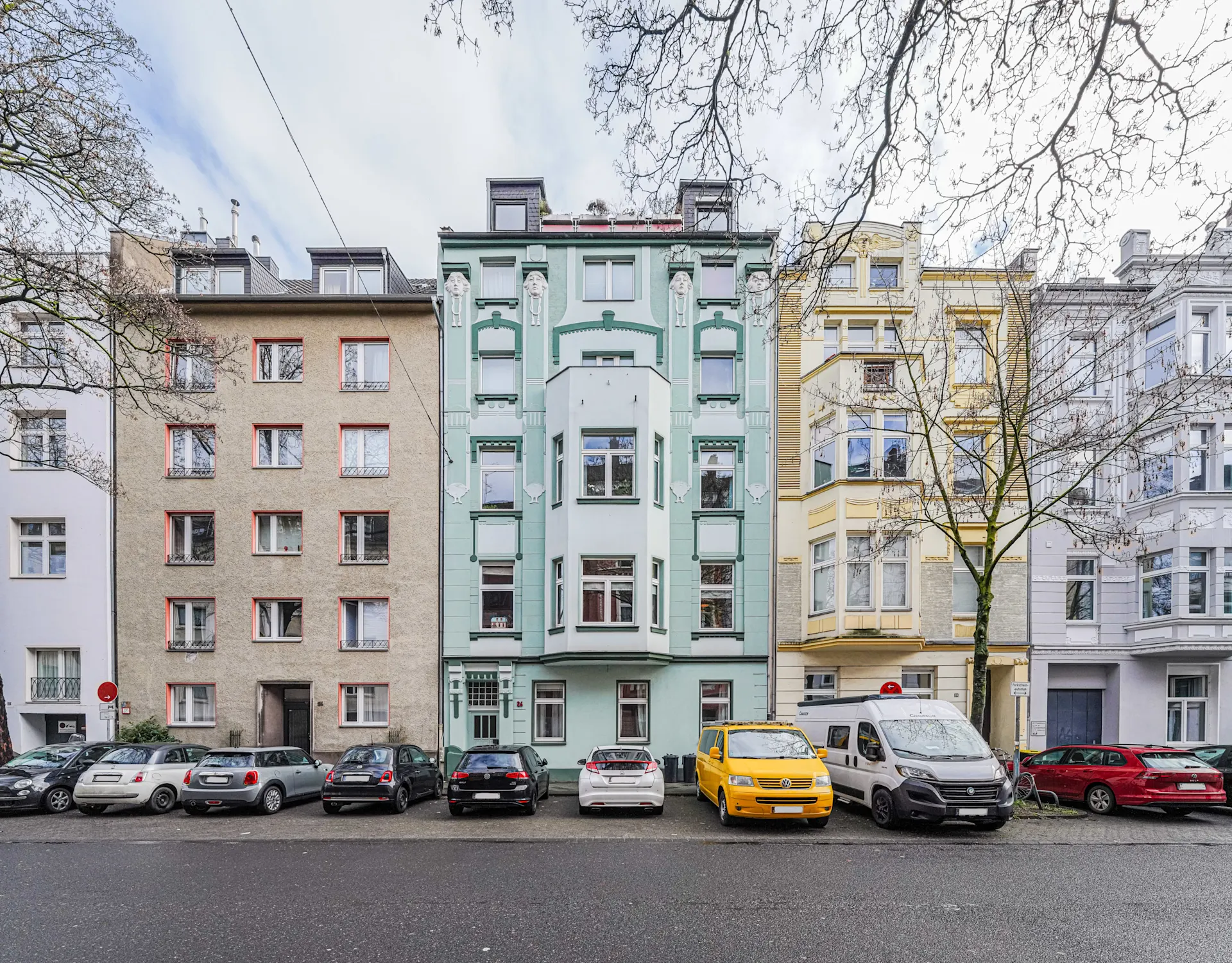 Mehrstöckige Wohnhäuser mit Autos auf der Straße davor, Bäume ohne Blätter am Rand.