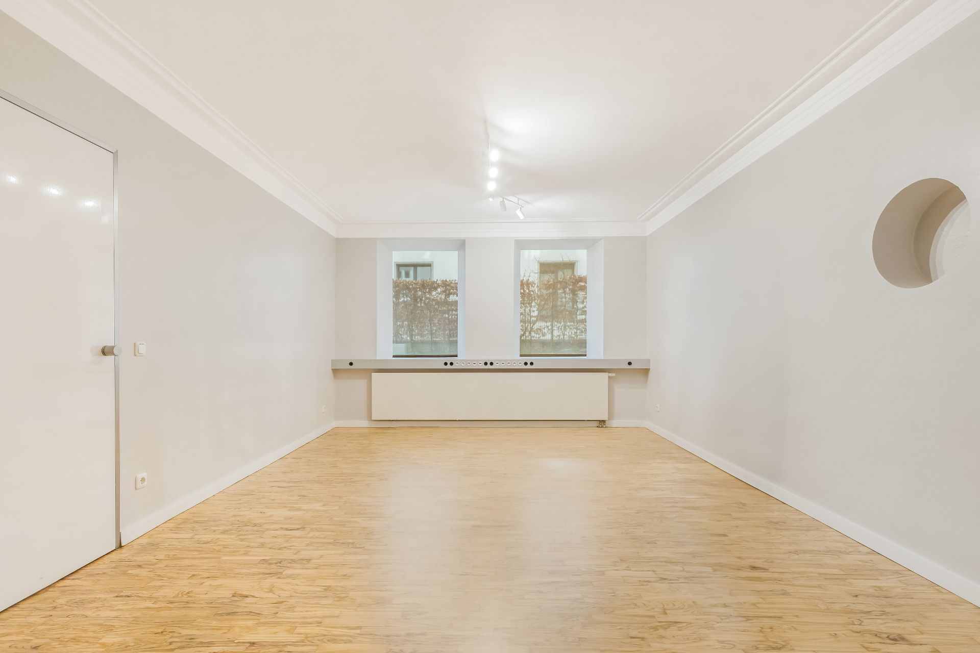 Heller, leerer Raum mit Holzfußboden, weißen Wänden und zwei Fenstern.