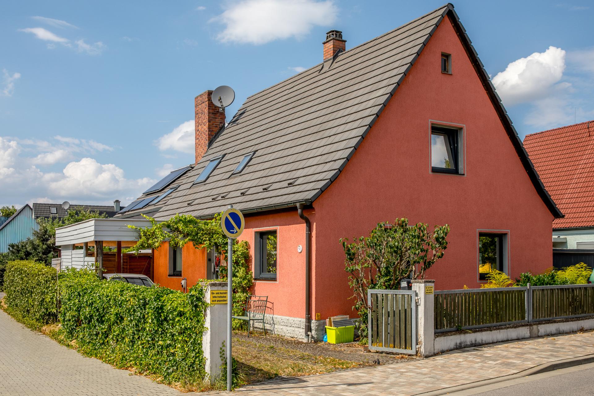 Rotes Einfamilienhaus mit Garten, Zaun und bepflanztem Vorgarten an einer ruhigen Straße.