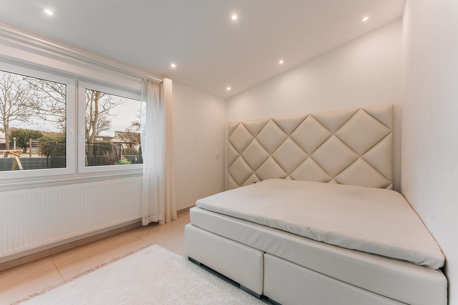 Helles Schlafzimmer mit großem Fenster, Doppelbett und gepolstertem Kopfteil.
