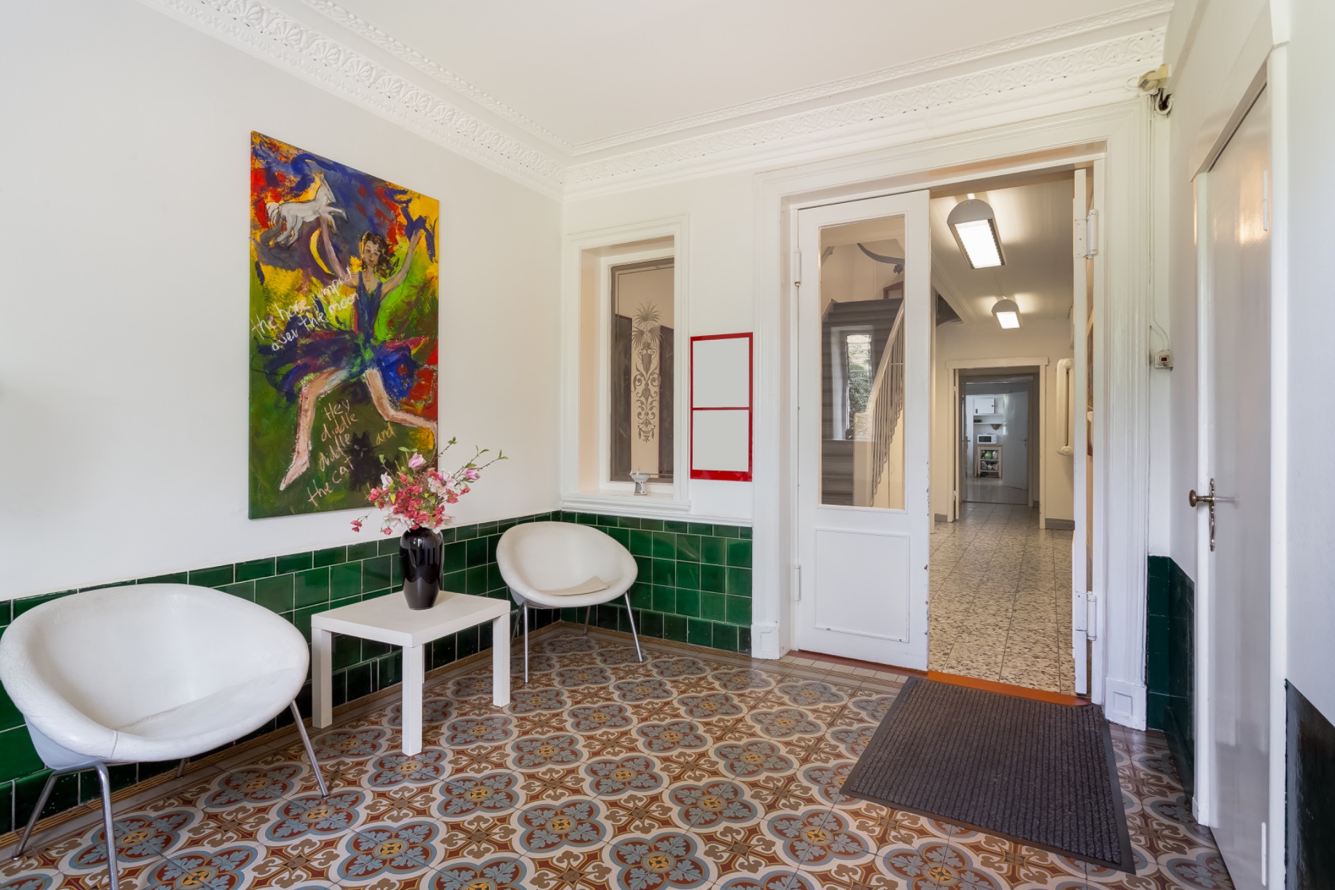 Heller Flur mit zwei weißen Stühlen, Tisch, Blumen und buntem Gemälde an der Wand.