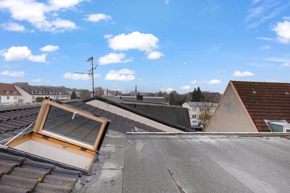 Blick von einem Wohnhausdach mit offenem Dachfenster auf umliegende Dächer und blauen Himmel.