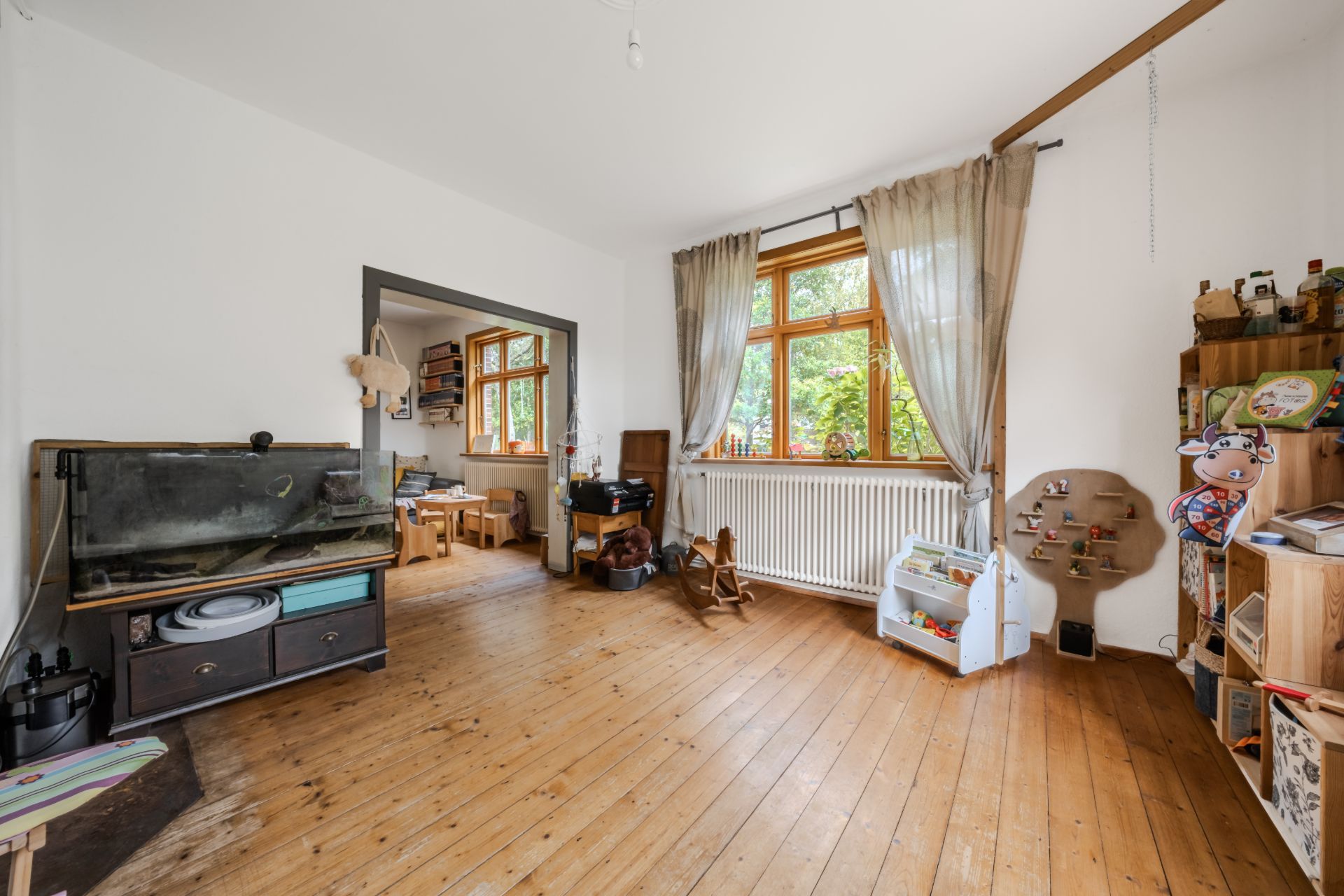 Helles Wohnzimmer mit Holzboden, Spielzeugregalen, großem Fenster und Blick in ein weiteres Zimmer.