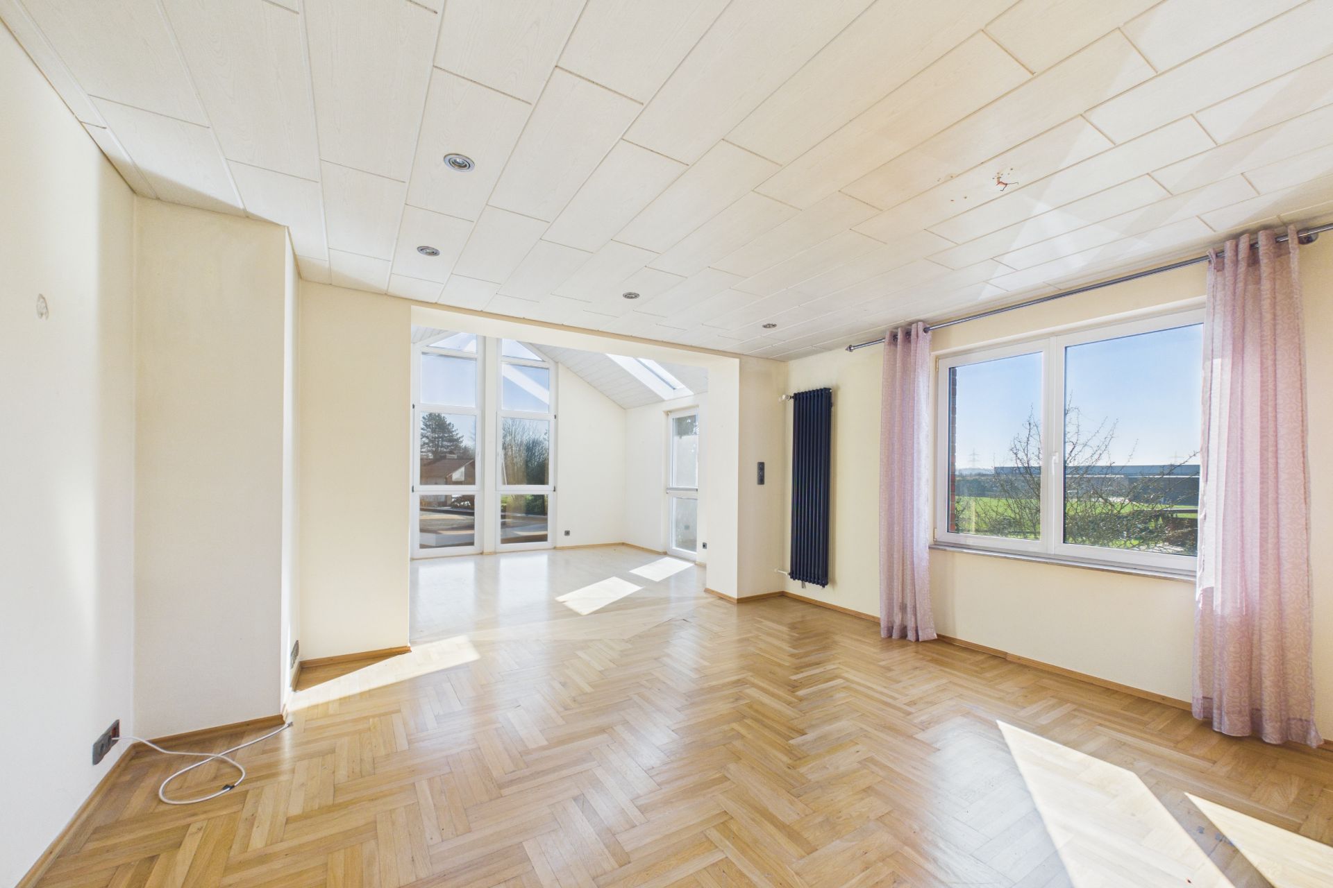 Helles Wohnzimmer mit Parkettboden, großen Fenstern und hellen Vorhängen.