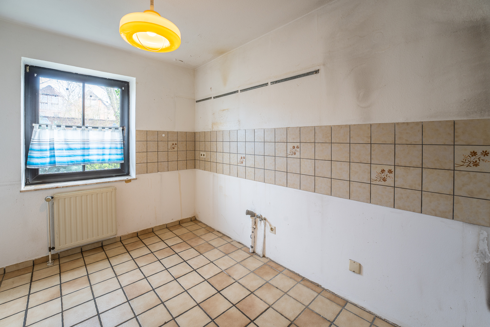 Leerer Raum mit gefliestem Boden, Fliesenspiegel und Fenster mit blau-weißem Vorhang.